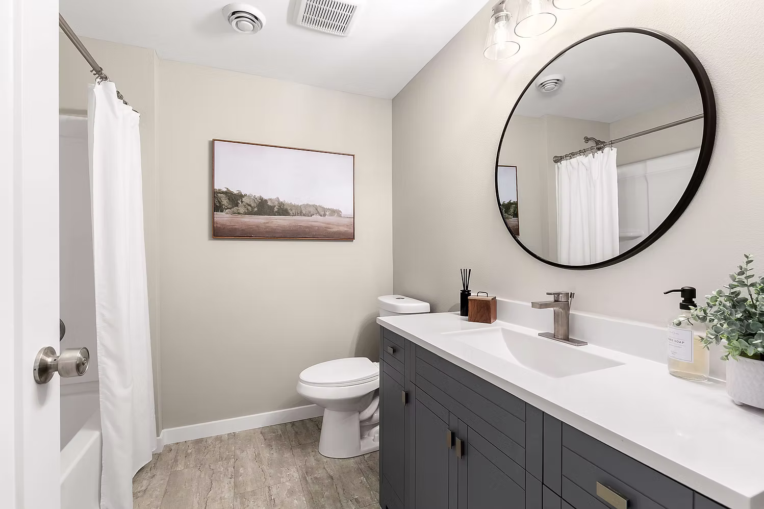 Gray shaker vanity with round mirror, white quartz top, and shower curtain